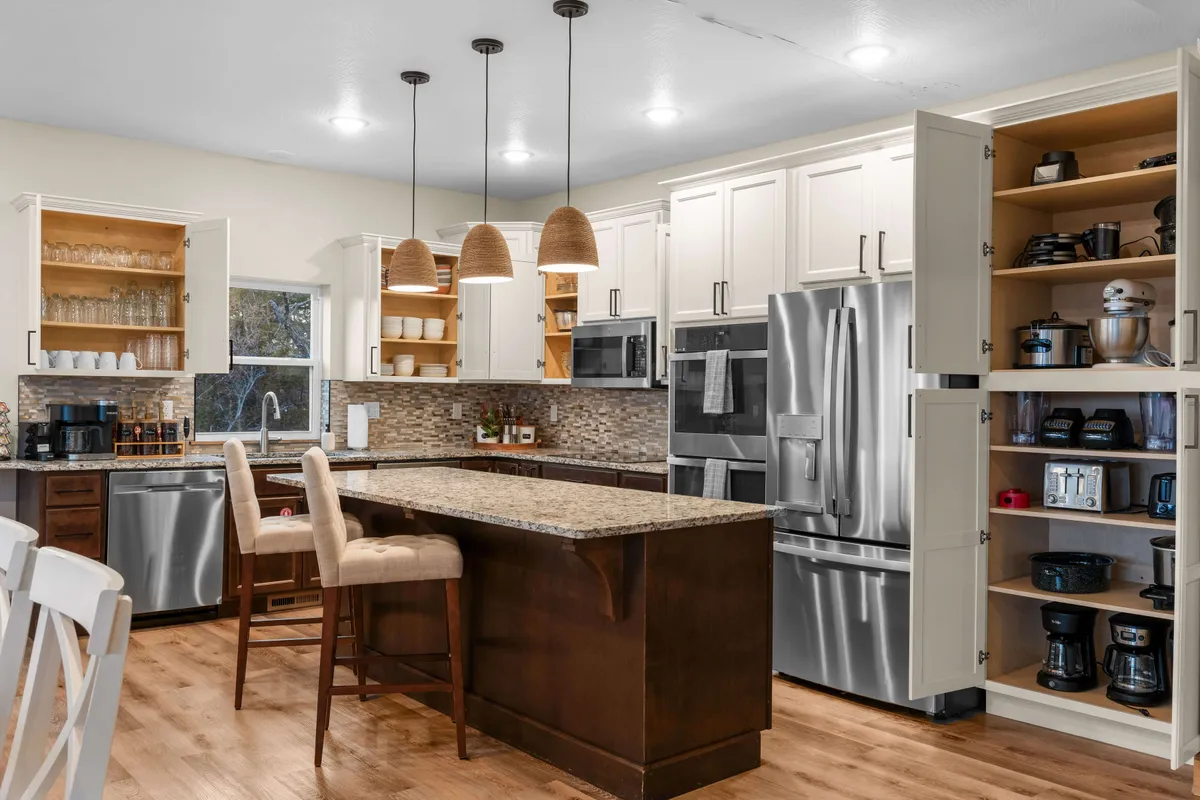 A bright, modern kitchen with a spacious island, stainless steel appliances, open shelving with glassware, and stylish pendant lights. The layout encourages group cooking and socializing, with plenty of prep space and gathering areas.