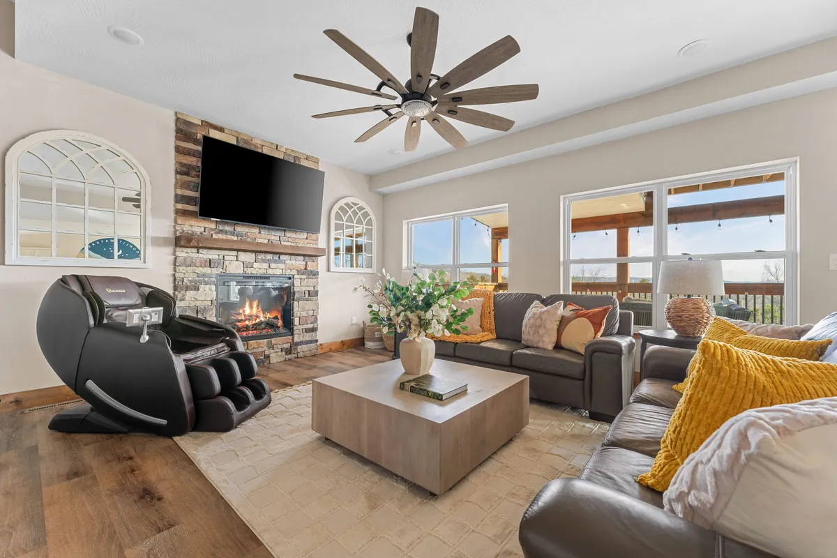 A welcoming living room featuring a glowing fireplace as the centerpiece, flanked by large windows that fill the space with natural light. Plush sofas with decorative pillows are arranged for conversation and relaxation, while a coffee table anchors the seating area, creating a homely and inviting atmosphere.
