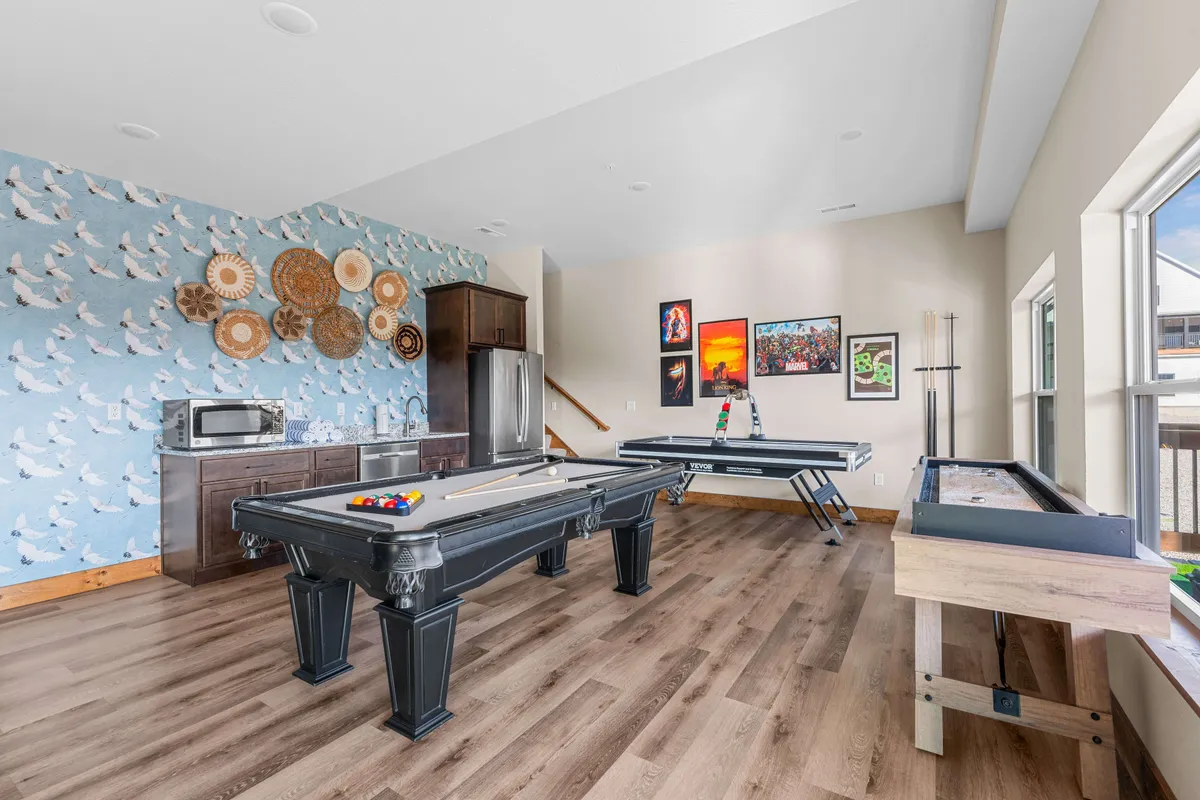 A recreational living room featuring a sleek pool table, decorative wall art, and a convenient kitchenette in the background. The space is set up for social interaction, games, and casual dining, with a relaxed and playful mood.