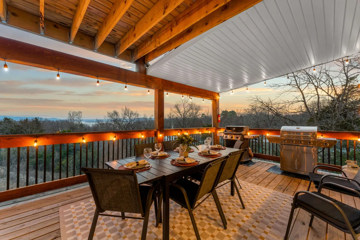 An inviting outdoor dining area equipped with a barbecue grill, a large table, twinkling string lights, and a beautiful sunset view. The setting is cozy, ideal for group meals, evening celebrations, and soaking in the outdoors.