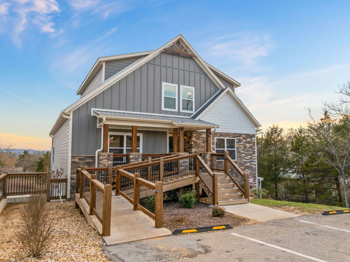 The image showcases the grand exterior of a spacious single-family house surrounded by nature in Hollister, Missouri. The stone and siding facade, expansive deck area, and large windows create an inviting atmosphere. A wooden ramp leads to the entrance, hinting at easy access for all. The deck offers ample space for gathering, relaxing, or taking in scenic sunsets.