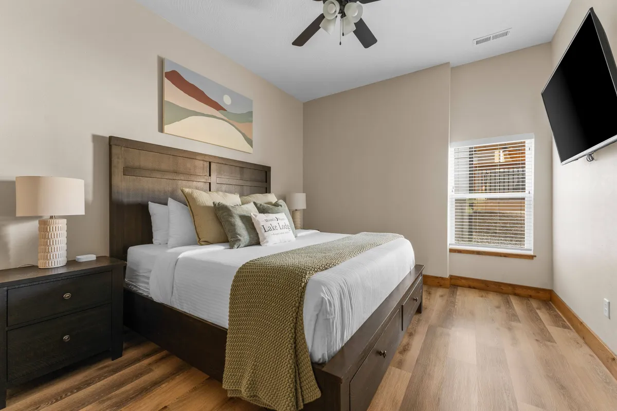 A tranquil bedroom with a plush bed, attractive artwork above the headboard, a nightstand, window, and a wall-mounted TV. The room is softly lit with natural light and exudes a serene, cozy atmosphere ideally suited for resting or watching TV.