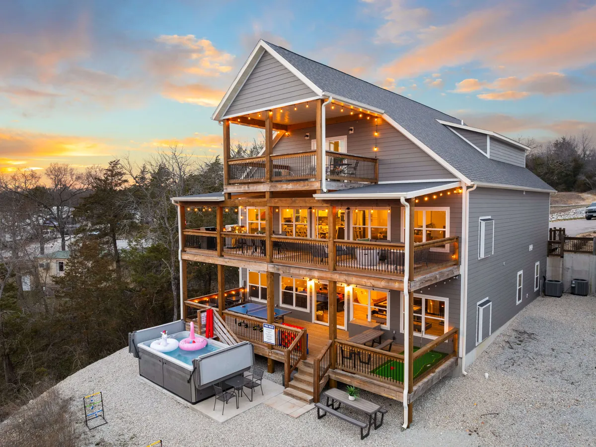 A vibrant exterior view featuring a spacious multi-level deck with ample outdoor seating, a hot tub, and festive string lights draped above. The sunset sky provides a warm backdrop, creating an inviting atmosphere ideal for group gatherings. The deck design suggests plenty of space for team activities and relaxation after a productive day.