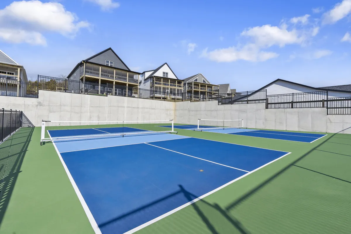 This image highlights lively tennis courts surrounded by neighboring houses and a clear blue sky. People can be seen engaged in tennis matches, with hints of social gatherings happening courtside. The scene conveys an energetic and active atmosphere, perfect for group sporting events or friendly competitions.